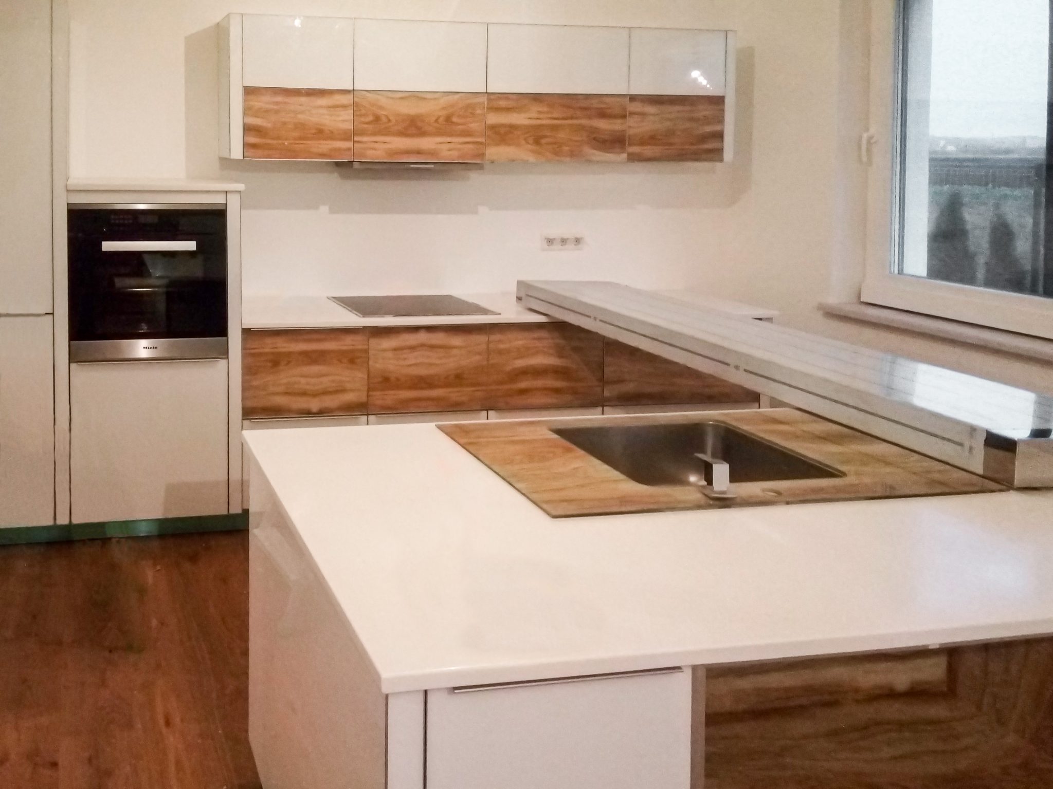 Perspectivă de ansamblu a unei bucătării albe cu insulă și blat Corian, cu mânere profil discrete și turn înalt cu cuptor încorporat.