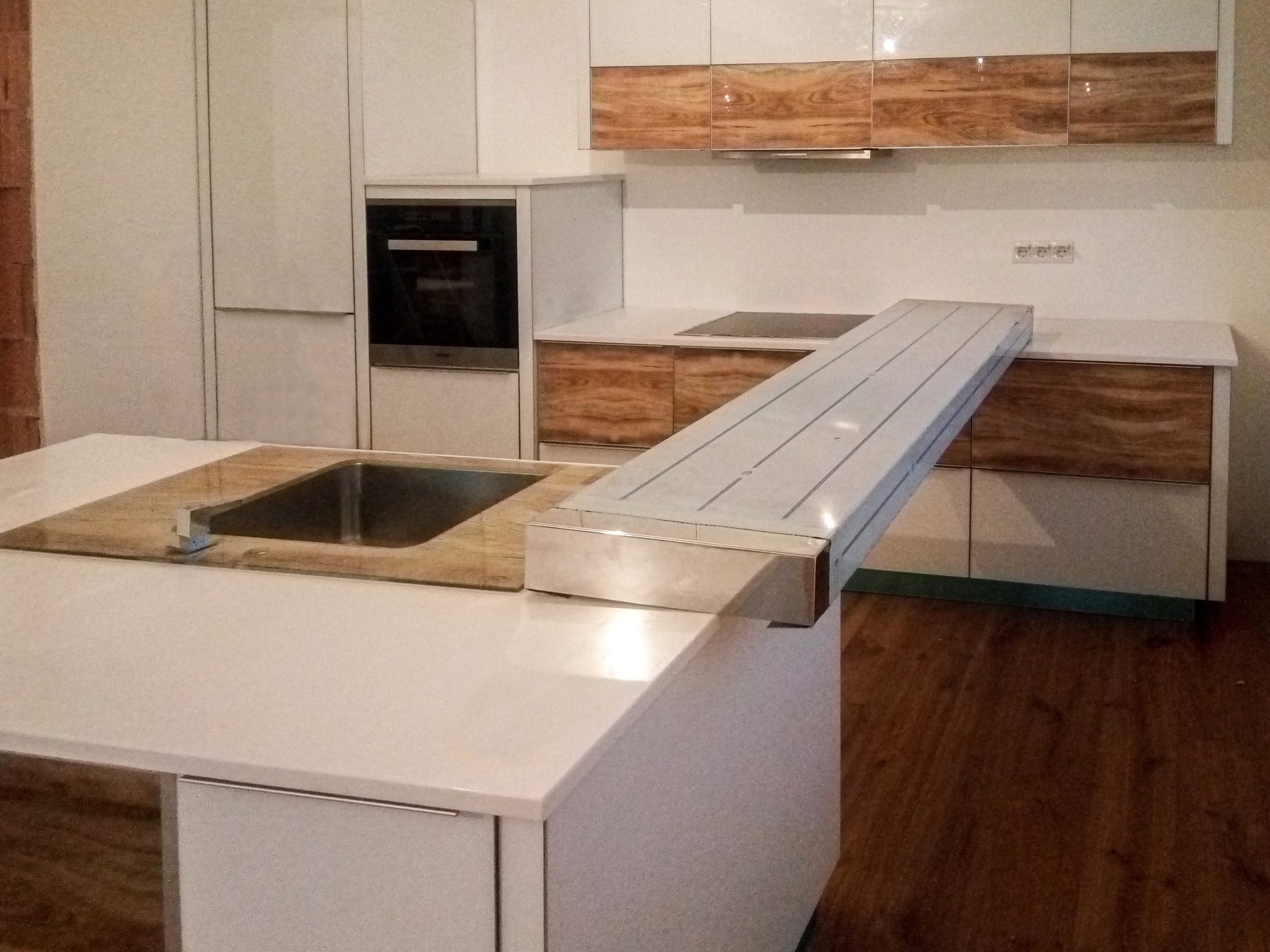 Perspectivă de ansamblu a unei bucătării albe cu insulă și blat Corian, cu mânere profil discrete și turn înalt cu cuptor încorporat.