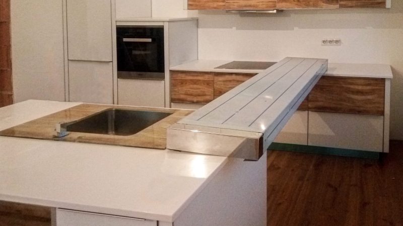 Perspectivă de ansamblu a unei bucătării albe cu insulă și blat Corian, cu mânere profil discrete și turn înalt cu cuptor încorporat.