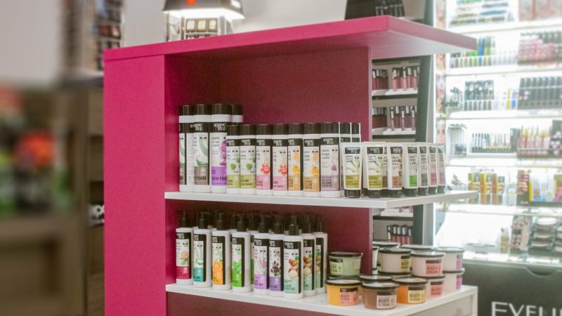 Stand comercial mobil pe roti cu rafturi fixe pentru produse cosmetice.