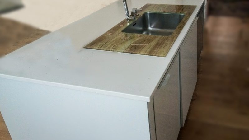 Prim-plan cu o chiuvetă modernă, încastrată într-un blat de sticlă securizată cu finisaj ce imită lemnul, parte dintr-o bucătărie cu blat Corian.